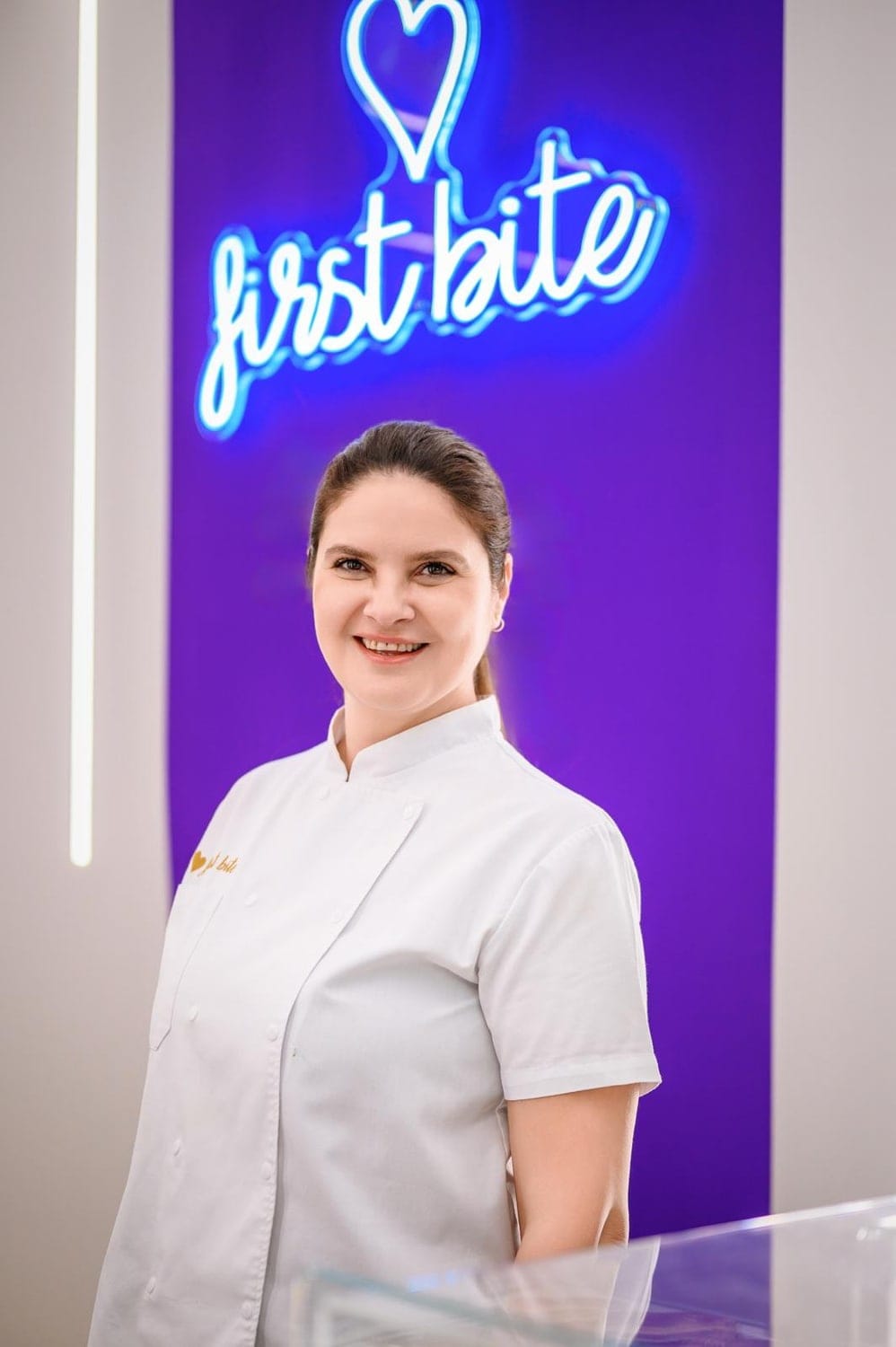 Femeie zâmbitoare în tunică albă de cofetar, în fața unui perete mov cu un neon „first bite”