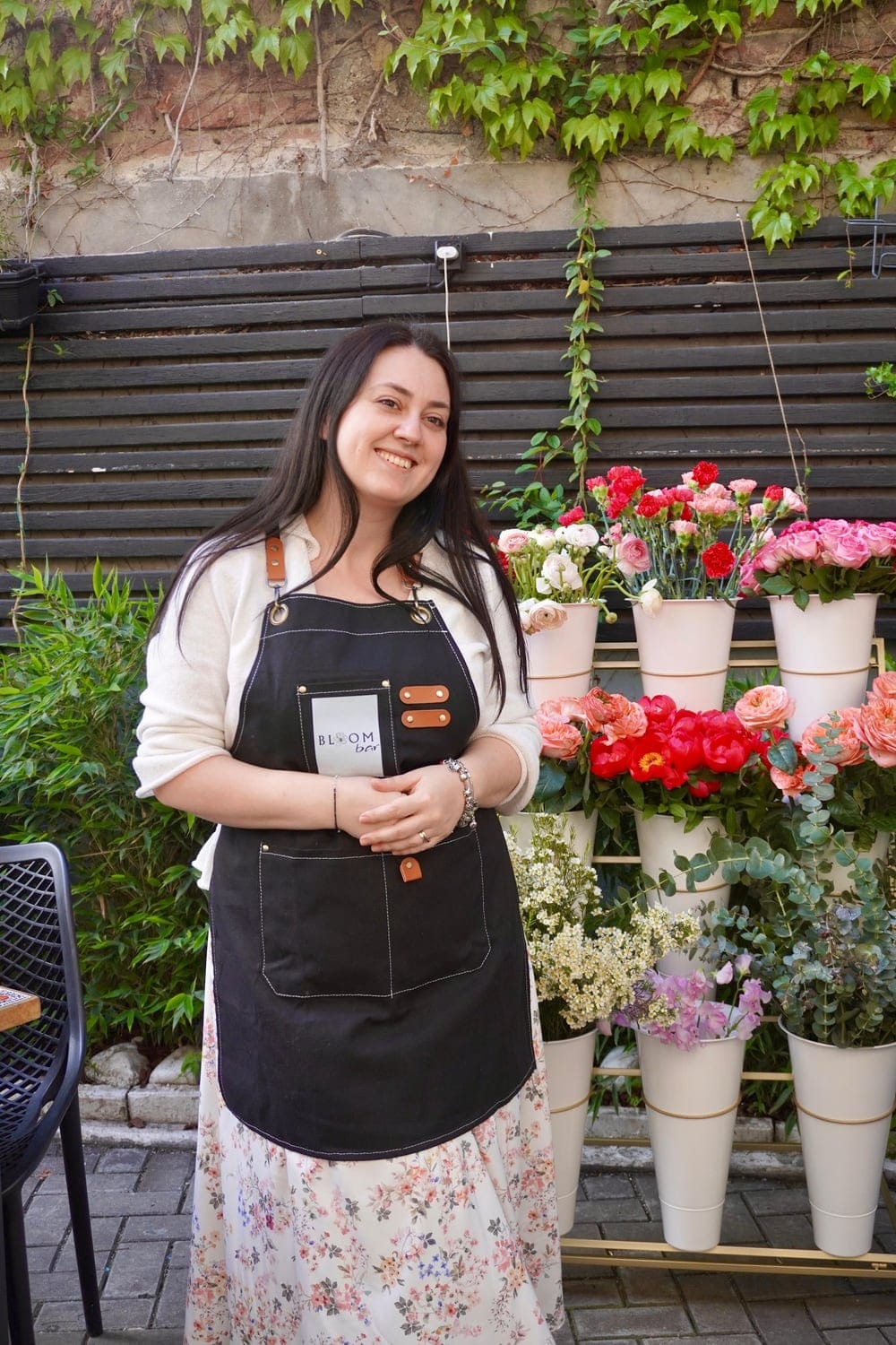 Florist cu șorț negru lângă un raft cu găleți albe pline cu trandafiri, bujori și garoafe