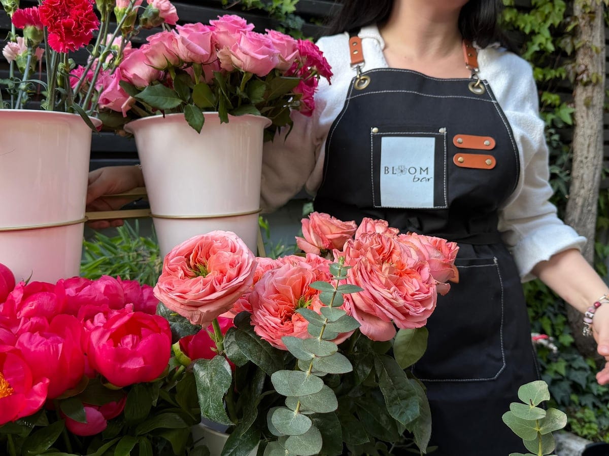 Bujori, trandafiri și eucalipt în găleți albe, lângă un șorț cu sigla BloomBar