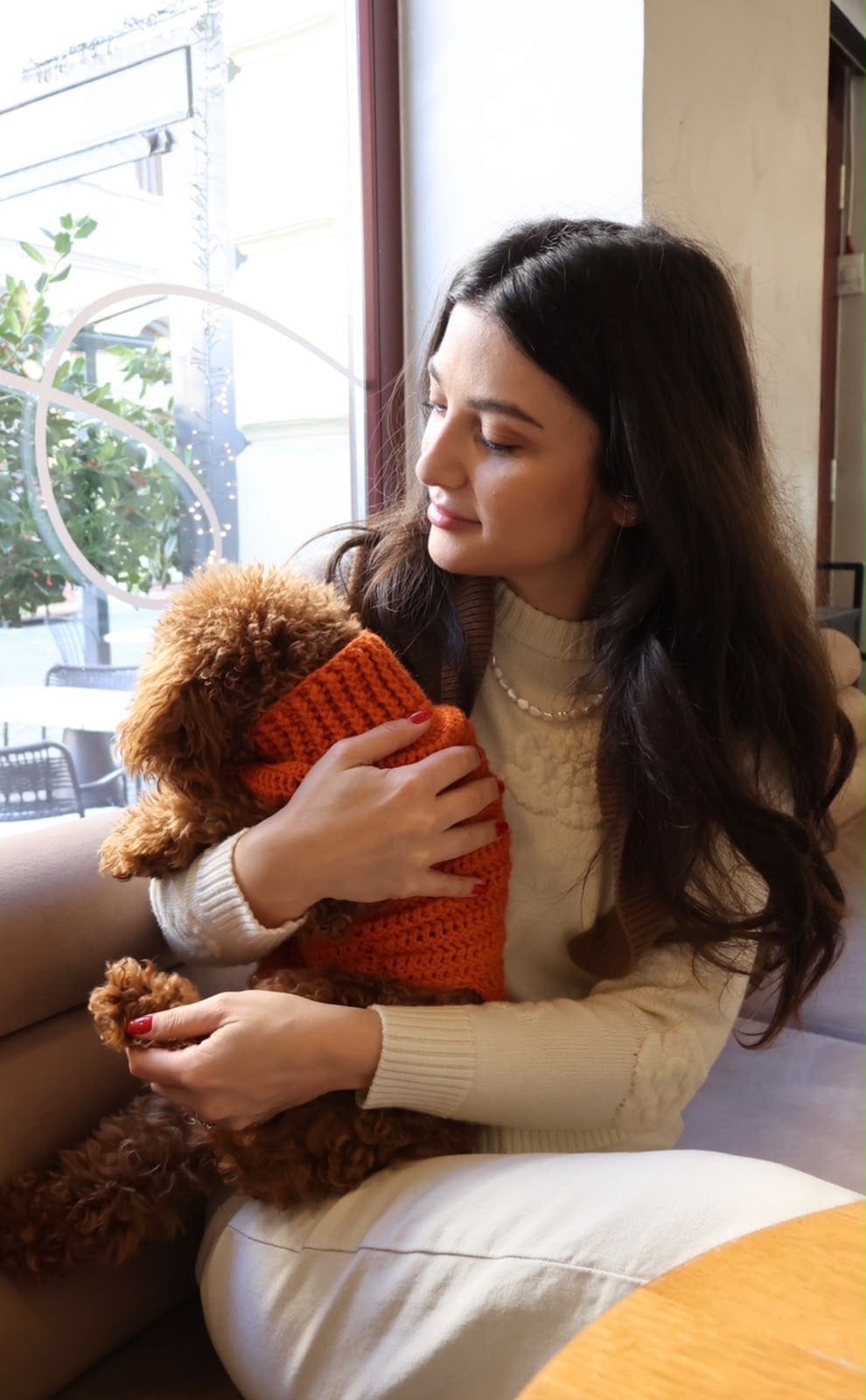 Tânără care ține în brațe un câine pudel îmbrăcat într-un pulover portocaliu, la fereastra unei cafenele