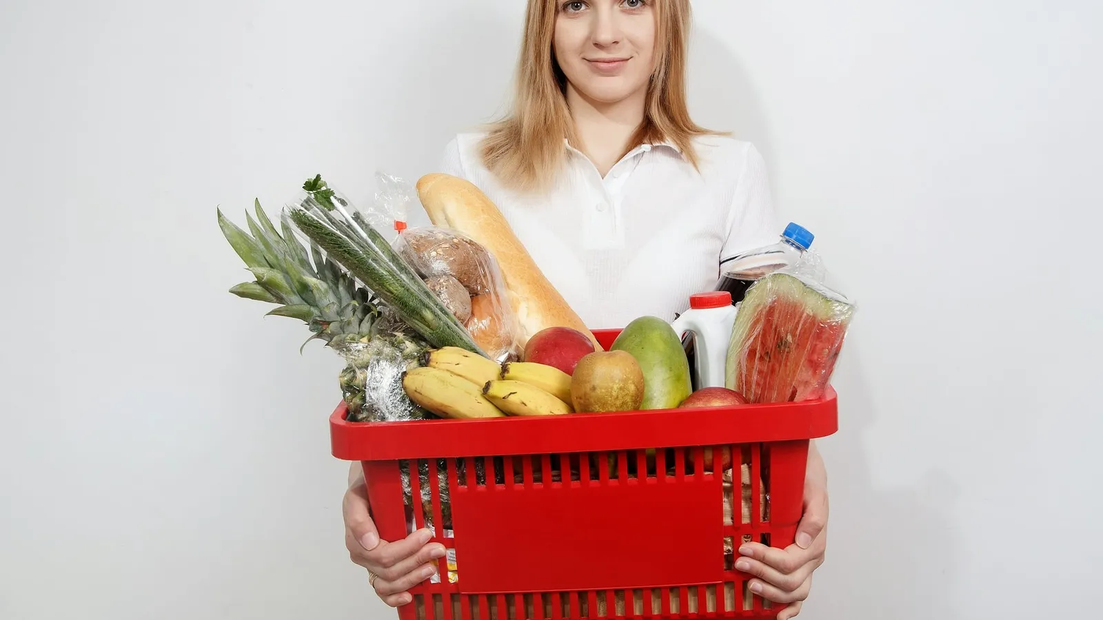 O femeie ține un coș roșu de cumpărături plin cu fructe, legume și pâine