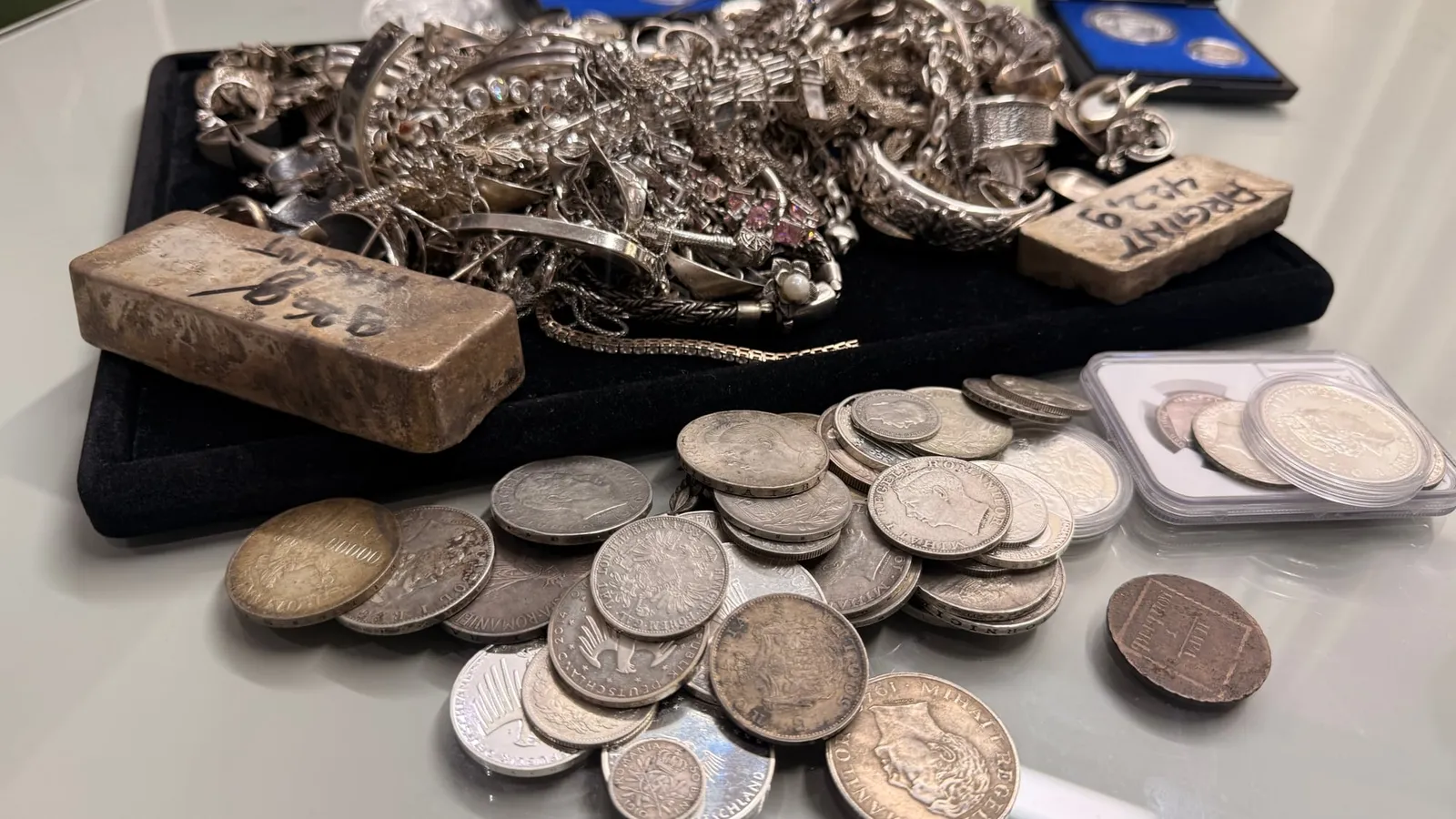 Bijuterii de argint, două lingouri și monede vechi de argint pe o tavă neagră, într-o casă de amanet
