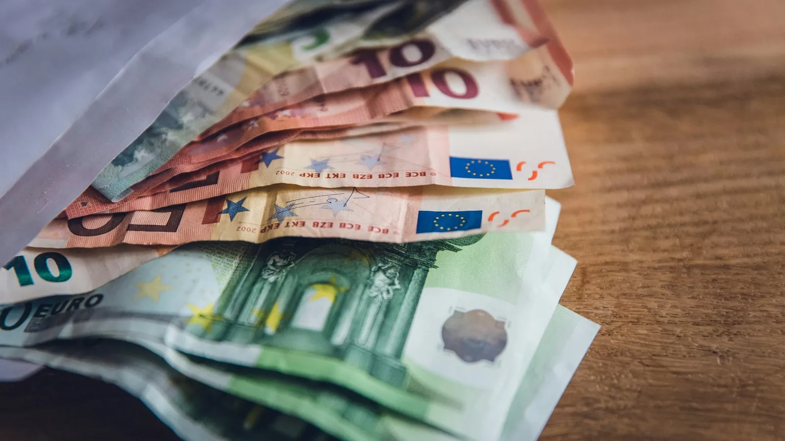 Bancnote euro de 10, 50 și 100 de euro ieșind dintr-un plic, pe o masă de lemn