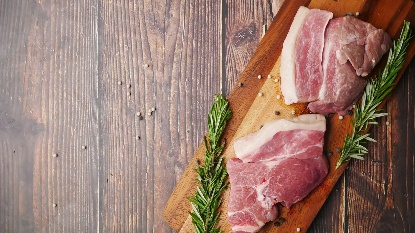 Bucăți de carne crudă de porc pe un tocător de lemn, cu rozmarin