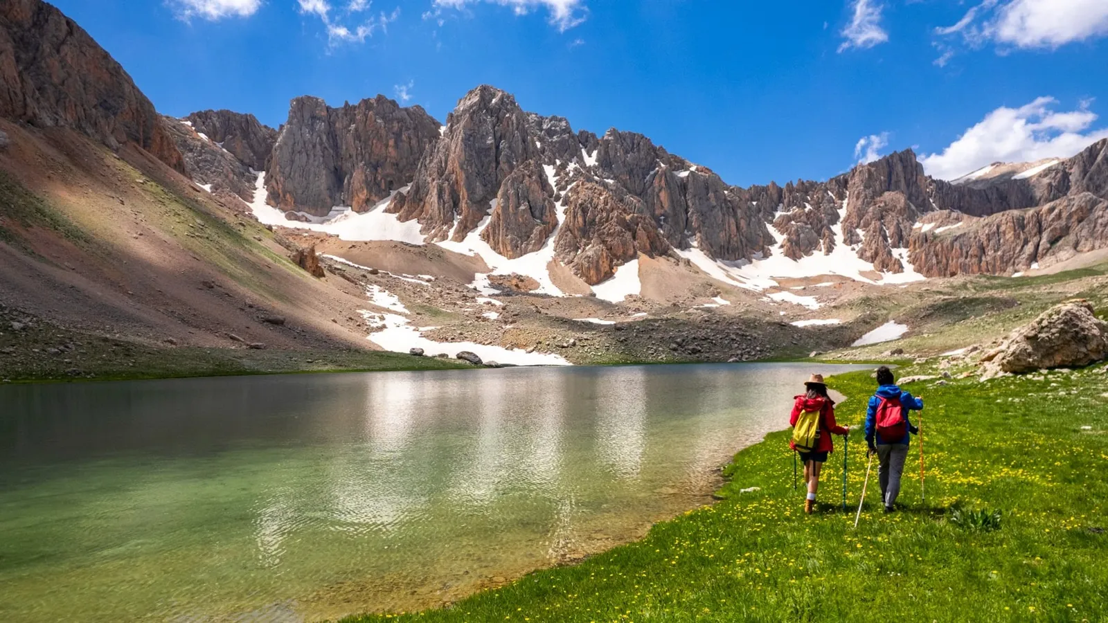 Doi drumeți merg pe malul unui lac de munte, cu vârfuri stâncoase acoperite parțial de zăpadă
