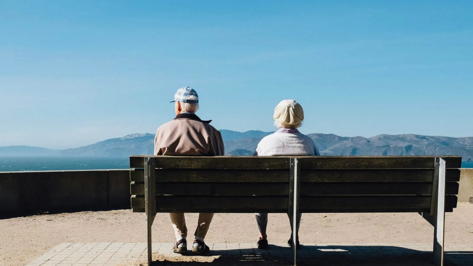 Doi pensionari stau pe o bancă, cu fața spre mare
