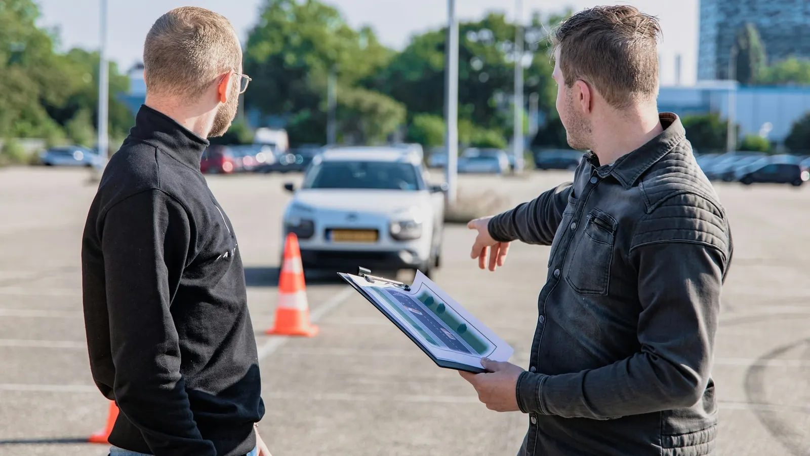 Instructor auto cu un clipboard arată spre o mașină pe un traseu marcat cu conuri, lângă un cursant