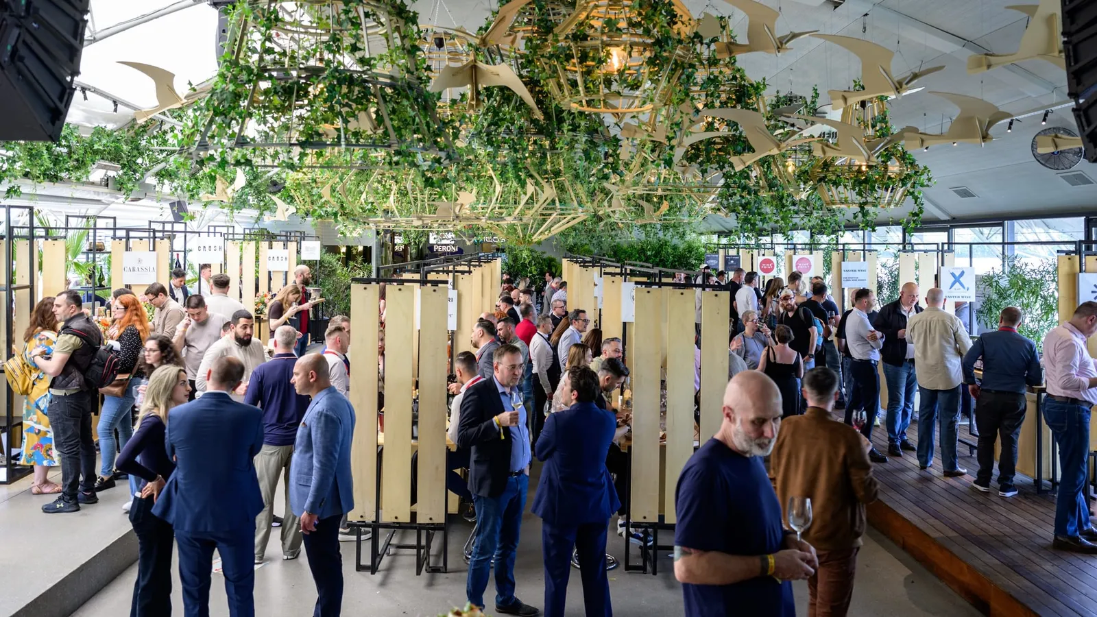 Vizitatori la standurile de vin dintr-o sală decorată cu plante agățătoare și păsări din lemn