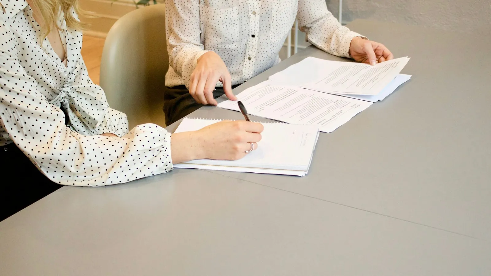 Două persoane la o masă, una semnează documente, cealaltă îi arată un contract
